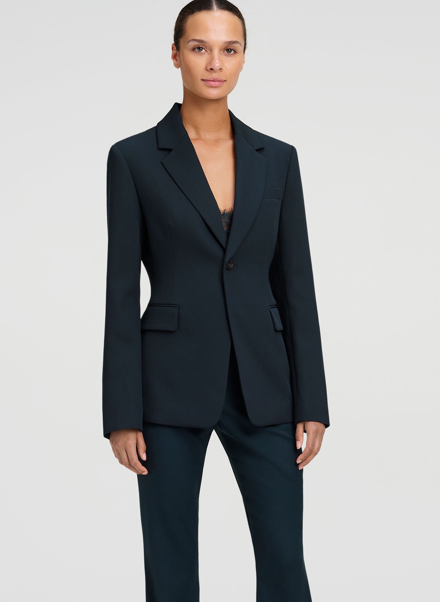 A model wearing a dark green tailored blazer with matching trousers, photographed against a light gray studio background.