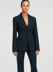 A model wearing a dark green tailored blazer with matching trousers, photographed against a light gray studio background.