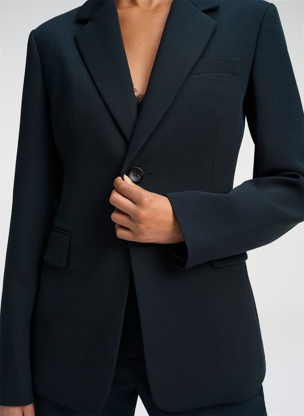 A close-up of a person wearing a dark green tailored blazer, with the hand adjusting the front button. The background is a clean, neutral light gray studio backdrop.