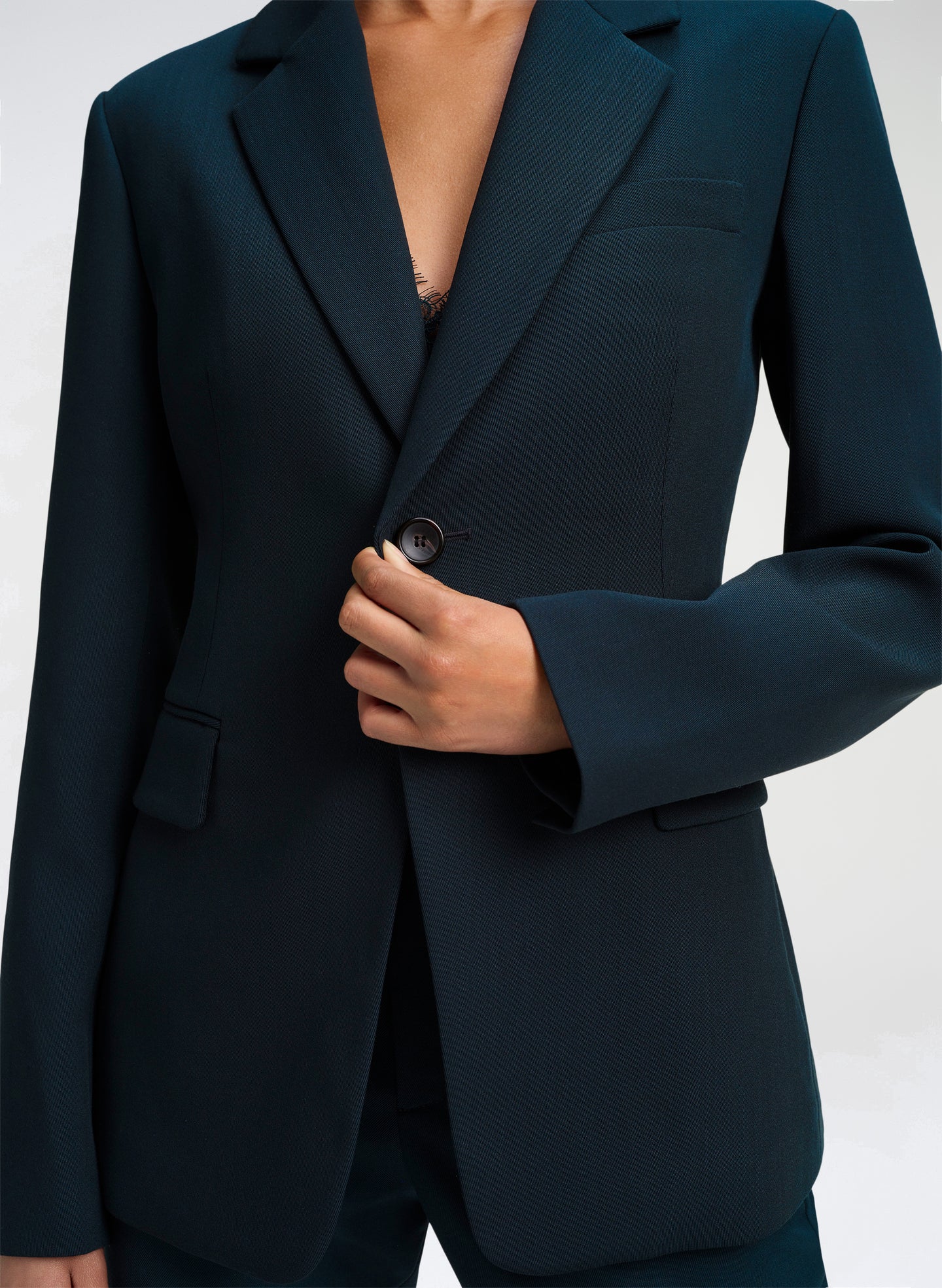 A close-up of a model wearing a dark green tailored blazer, showing the lapels, button closure, and hand adjusting the front.