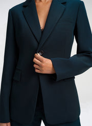 A close-up of a model wearing a dark green tailored blazer, showing the lapels, button closure, and hand adjusting the front.