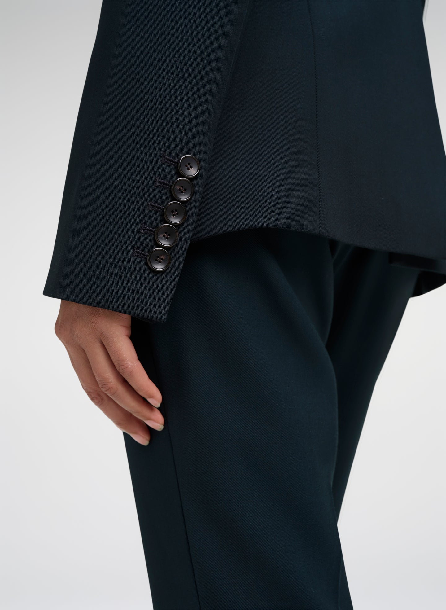 Close-up of a person wearing tailored dark green trousers and a matching blazer, showing the cuff with five buttons and part of the hand against a light gray studio background.