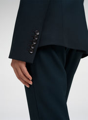 Close-up of a person wearing tailored dark green trousers and a matching blazer, showing the cuff with five buttons and part of the hand against a light gray studio background.
