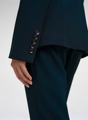 Close-up of a person wearing tailored dark teal trousers and a matching blazer, showing the back of the sleeve with five dark buttons and the hand resting near the hem.