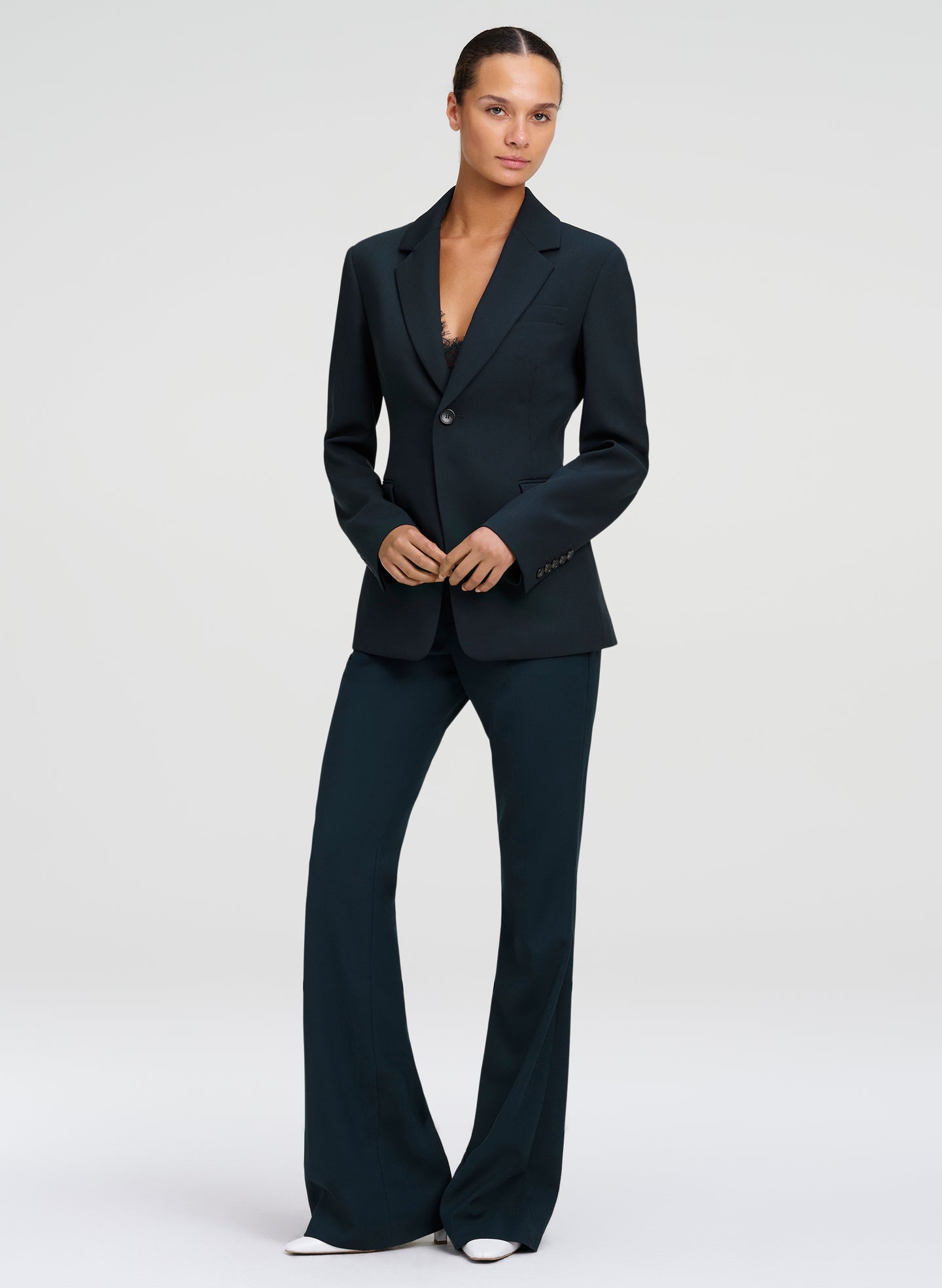 A model wearing a dark green tailored suit, consisting of a single-breasted blazer and matching wide-leg trousers, standing against a light gray studio background.