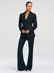 A model wearing a dark green tailored suit, consisting of a single-breasted blazer and matching wide-leg trousers, standing against a light gray studio background.