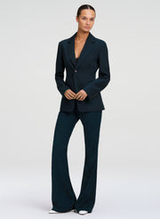 A model wearing a dark tealtwo-piece suit consisting of a single-breasted blazer and matching wide-leg trousers, paired with white pointed-toe heels, against a light gray studio background.