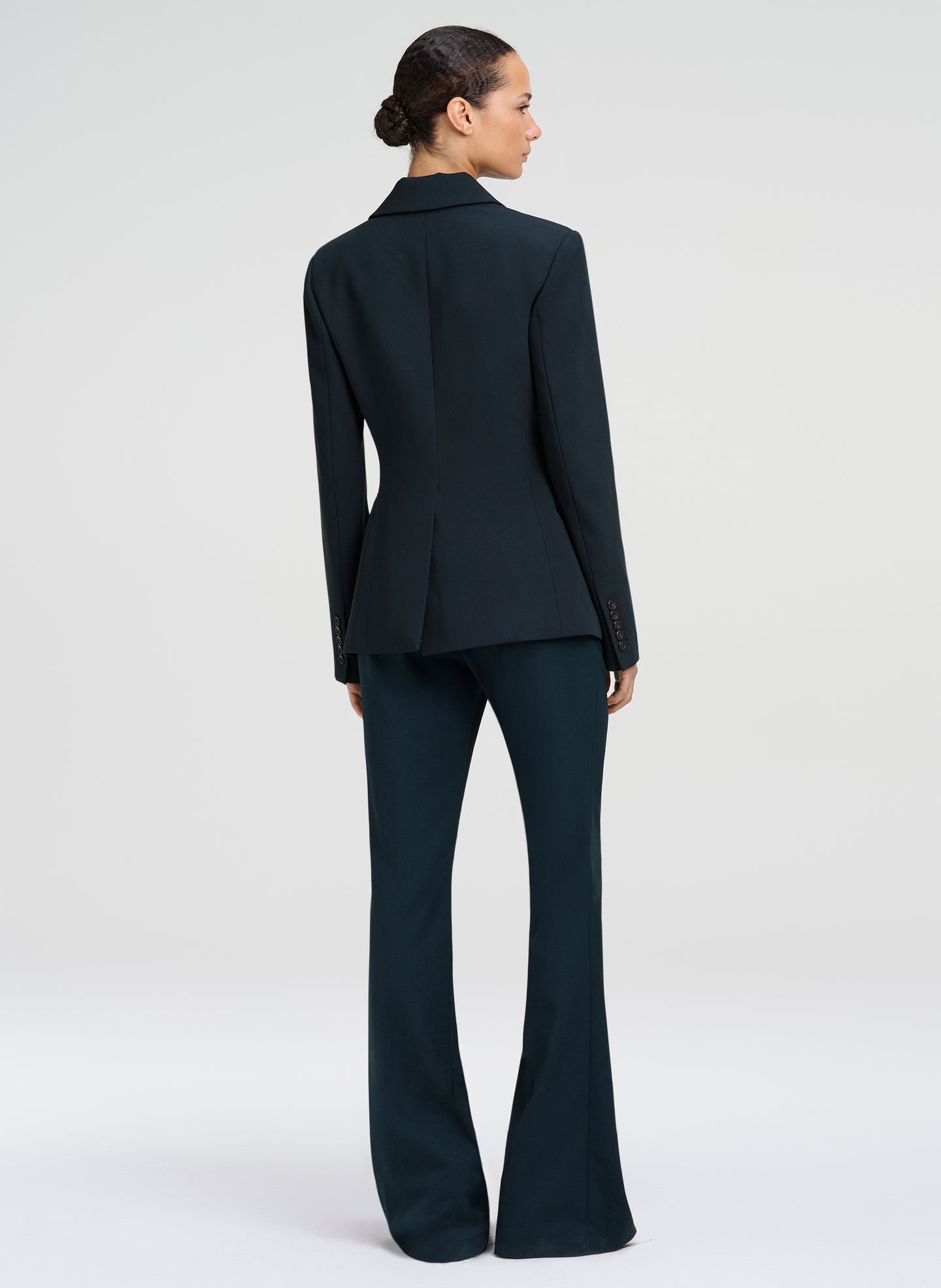 A model shown from the back wearing a dark green tailored suit, featuring a structured blazer with long sleeves and matching high-waisted flared trousers, set against a plain light gray studio background.
