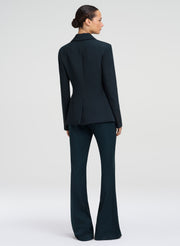 A model shown from the back wearing a dark green tailored suit, featuring a structured blazer with long sleeves and matching high-waisted flared trousers, set against a plain light gray studio background.