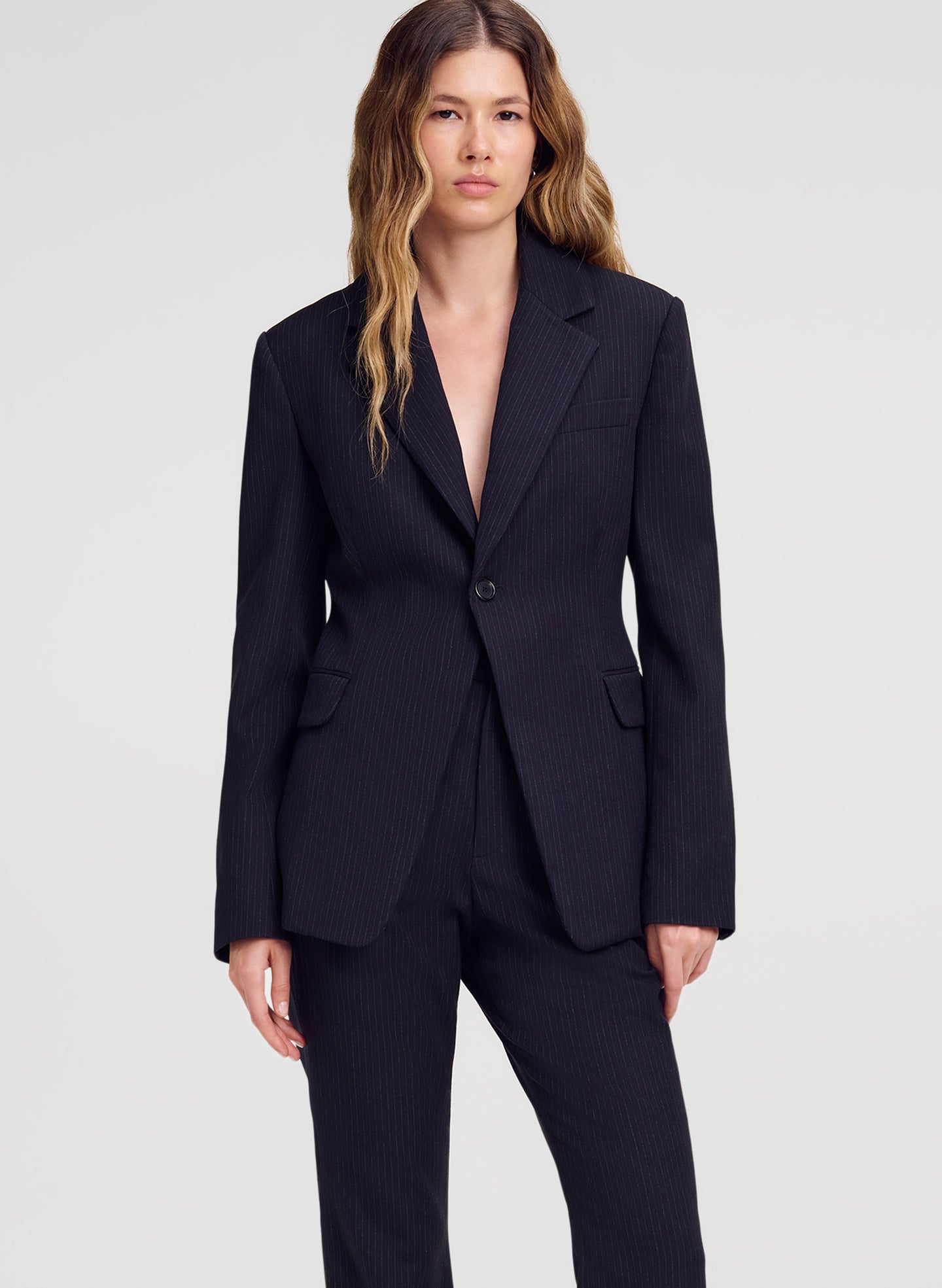 A woman modeling a dark navy pinstripe blazer with matching trousers against a light gray studio background.