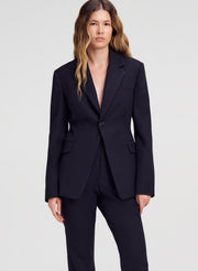 A woman modeling a dark navy pinstripe blazer with matching trousers against a light gray studio background.