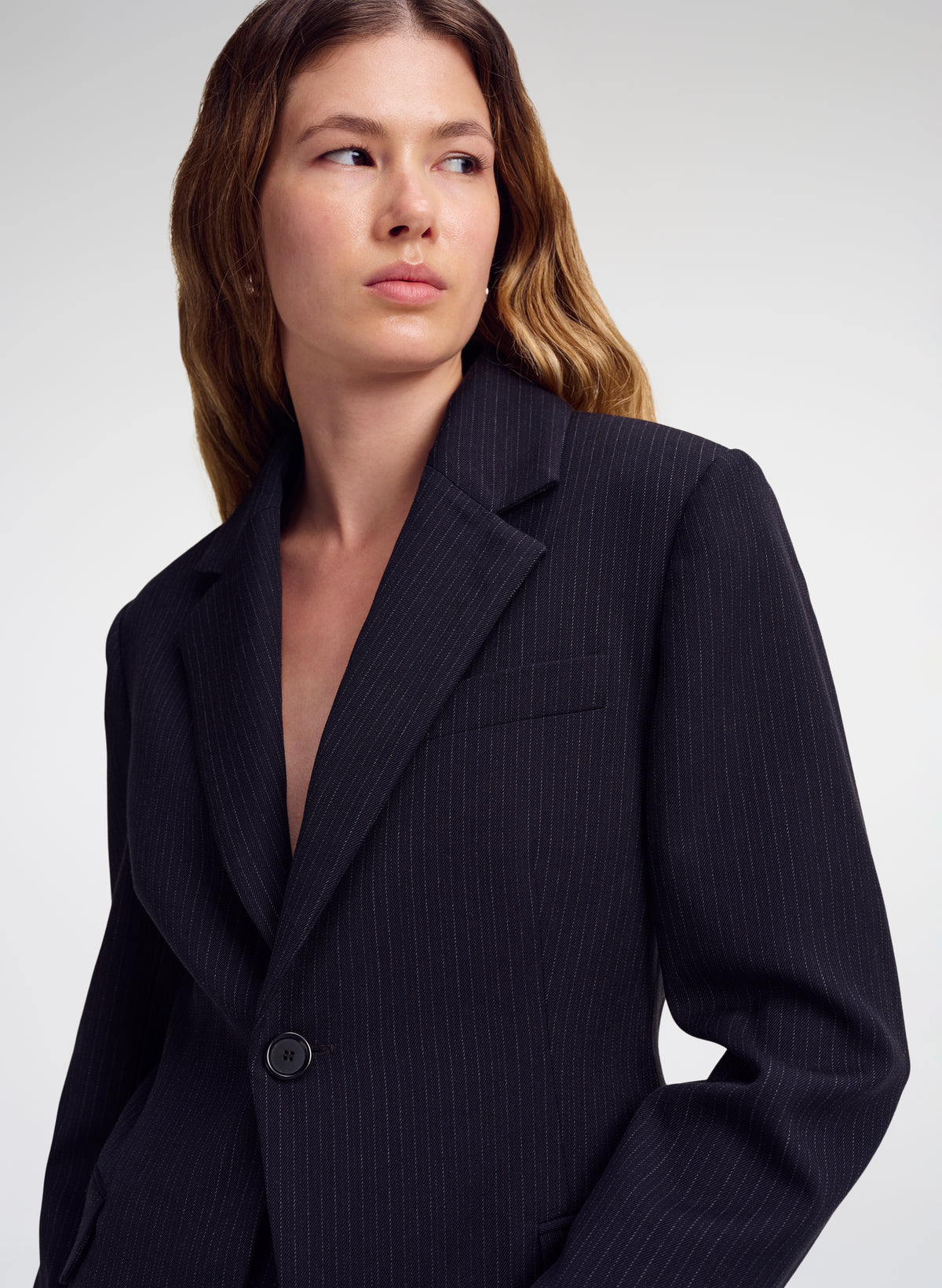 A woman wearing a dark navy pinstripe blazer, photographed from the chest up against a light gray studio background.