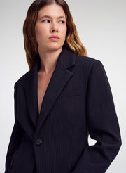 A woman wearing a dark navy pinstripe blazer, photographed from the chest up against a light gray studio background.