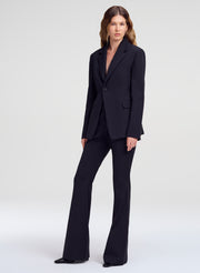 A model wearing a dark navy blue tailored suit with a single-breasted blazer and matching wide-leg flared trousers, standing against a light gray studio background.