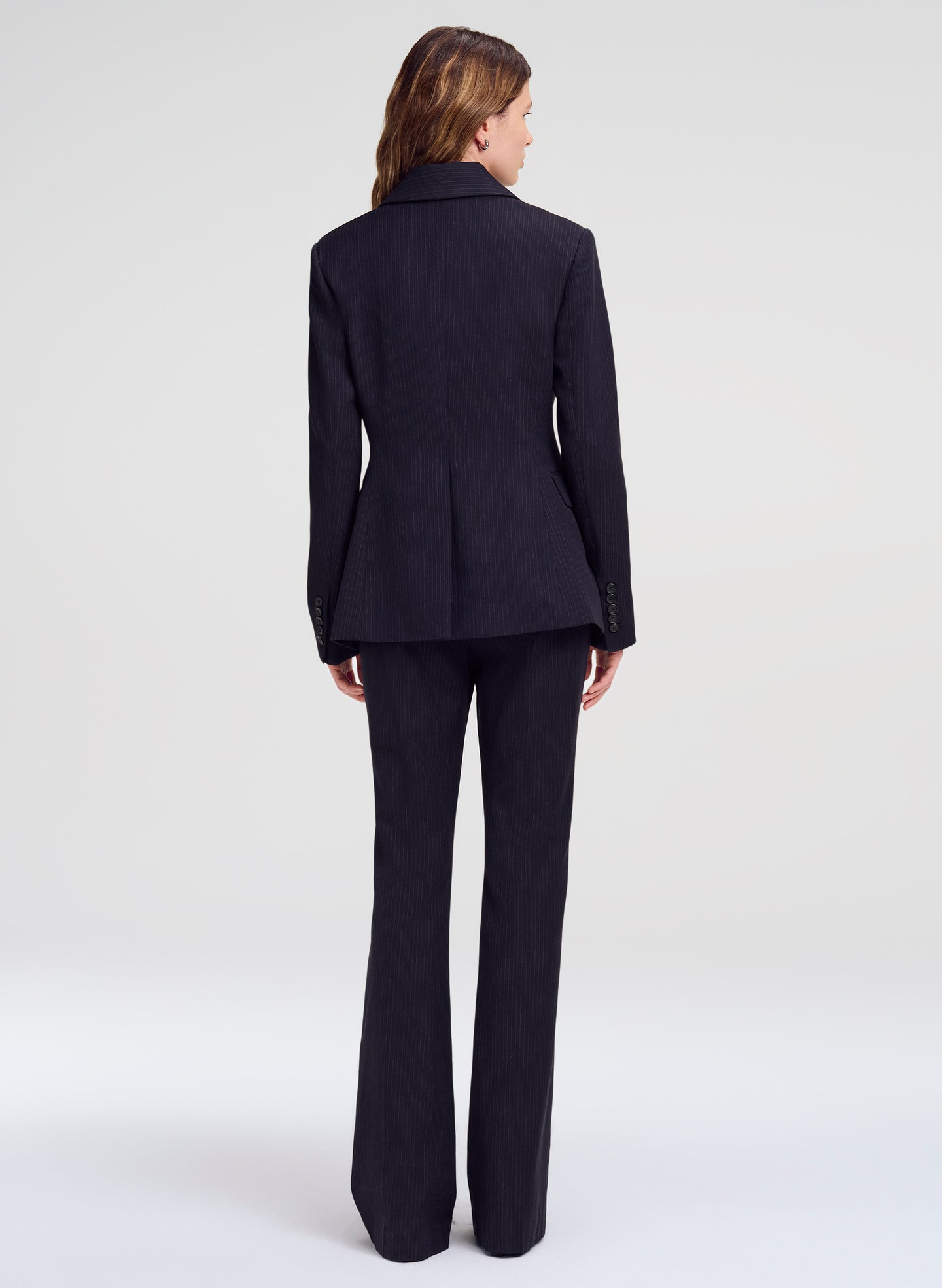 A woman viewed from the back wearing a dark navy tailored suit with a structured blazer and matching wide-leg trousers, photographed against a clean light gray studio background.