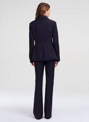A woman viewed from the back wearing a dark navy tailored suit with a structured blazer and matching wide-leg trousers, photographed against a clean light gray studio background.