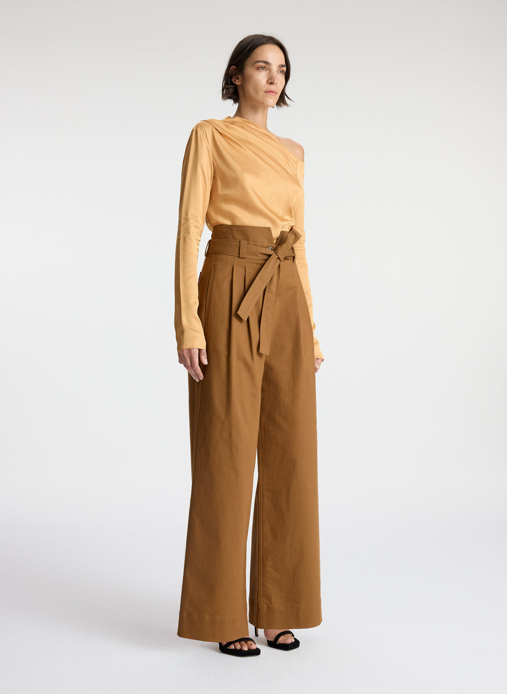 side view of woman wearing tan one shoulder long sleeve top and brown wide leg pants