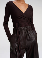 A close-up of a model wearing a dark brown long-sleeve wrap top with a matching tie belt and high-waisted brown leather trousers against a light gray studio background.