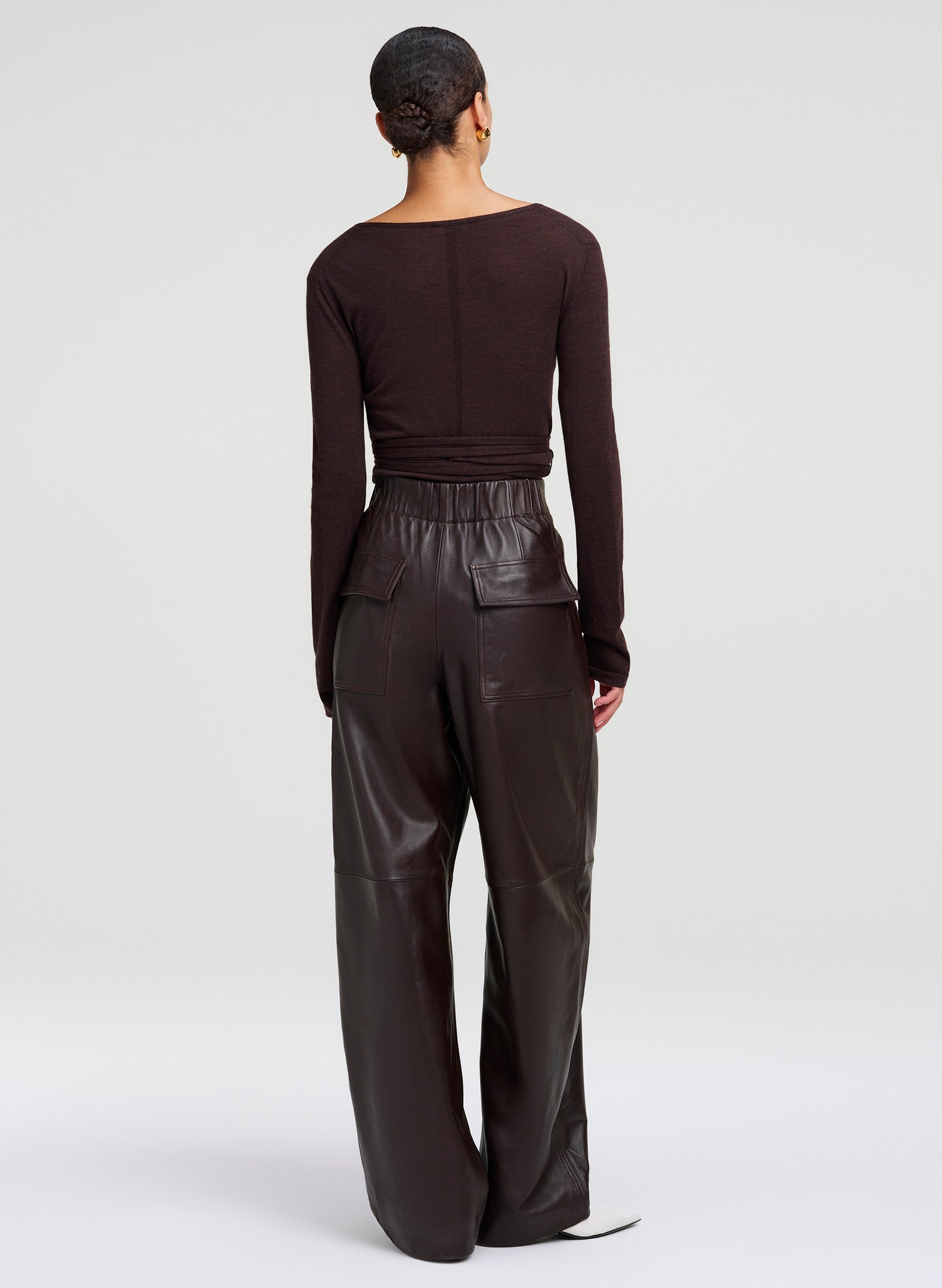 A model shown from the back wearing a dark brown long-sleeve top and matching dark brown leather wide-leg pants, set against a plain light gray studio background.