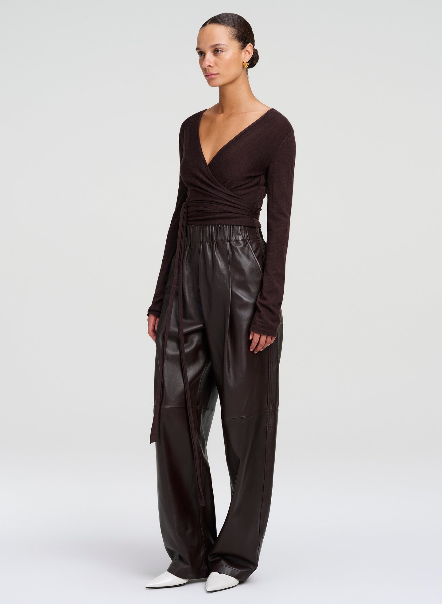 A model wearing a dark brown long-sleeve wrap-style top and matching wide-leg  leather pants, paired with white pointed shoes, against a light gray studio background.