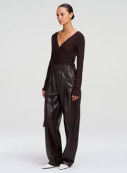 A model wearing a dark brown long-sleeve wrap-style top and matching wide-leg  leather pants, paired with white pointed shoes, against a light gray studio background.