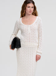 A woman modeling a cream-colored cable-knit two-piece set consisting of a long-sleeve henley top and a matching midi skirt, holding a black clutch bag against a light gray studio background.