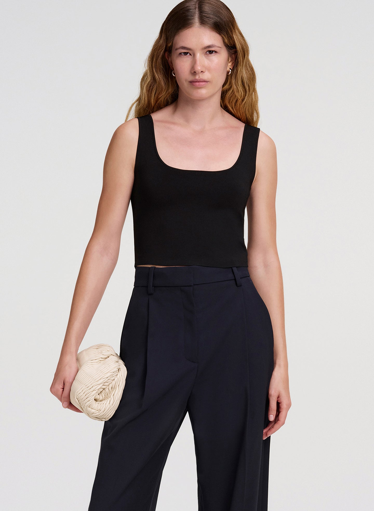 Woman wearing a black sleeveless top and dark pants, holding a beige clutch against a white background
