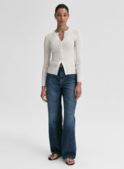 Woman wearing a white ribbed cardigan and blue jeans on a light gray background