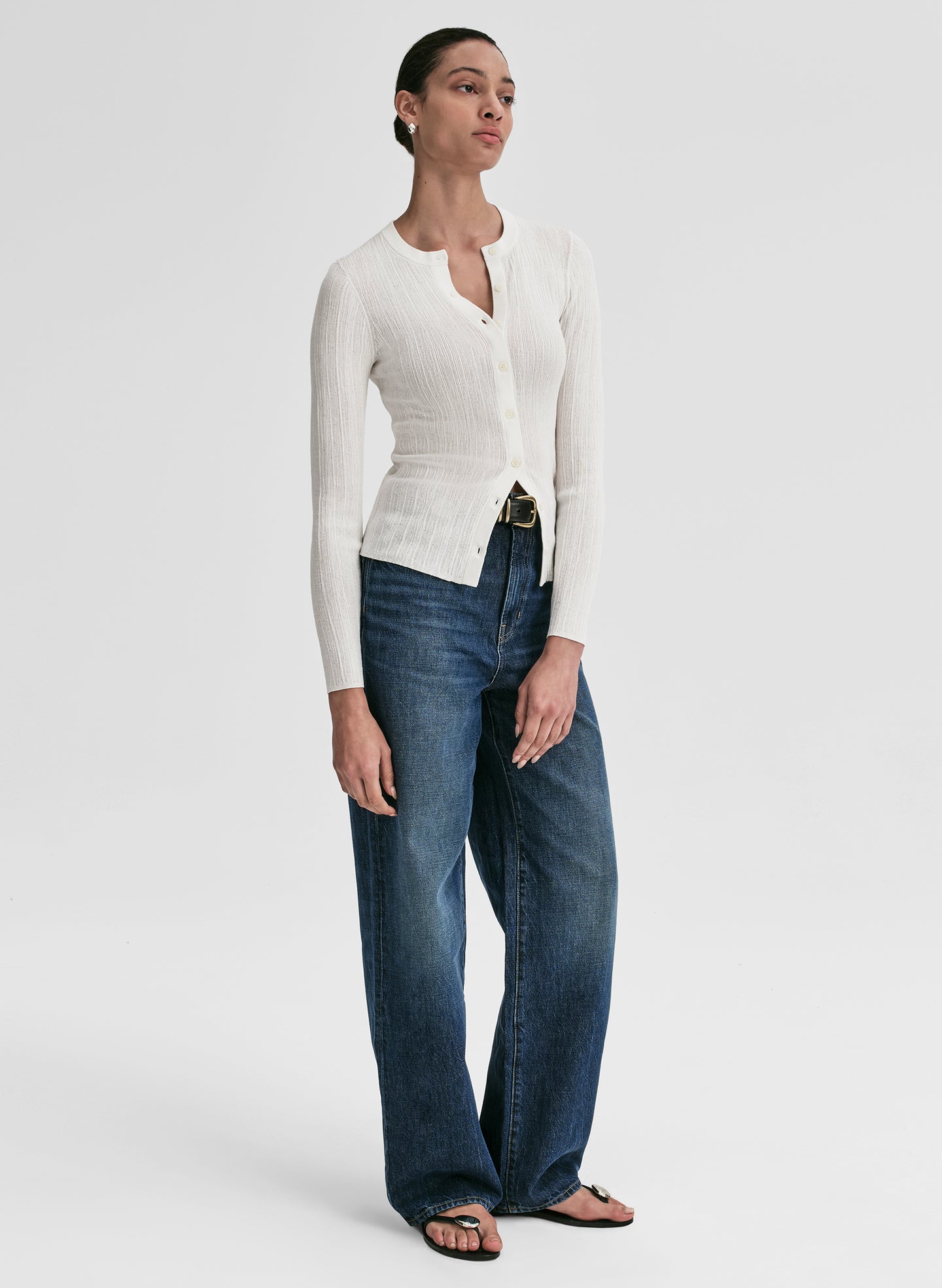 Woman wearing a white ribbed cardigan and blue jeans on a light gray background