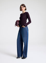 Woman wearing a dark purple long-sleeve top and blue jeans, holding a patterned handbag on a white background.