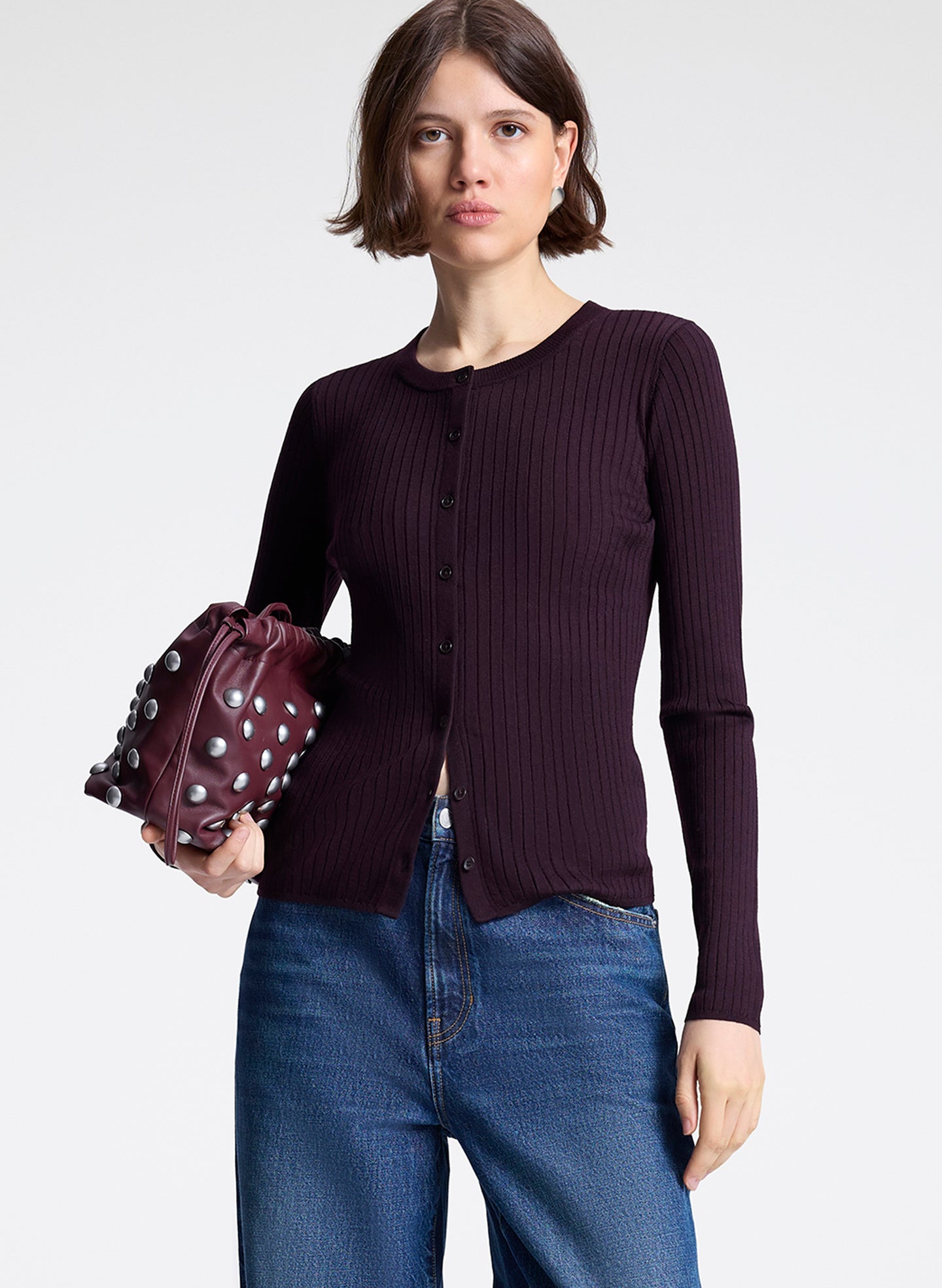 Woman wearing a dark purple ribbed sweater and blue jeans, holding a matching purple bag on a white background