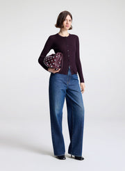 Woman wearing a dark purple cardigan and blue jeans holding a purple clutch against a white background