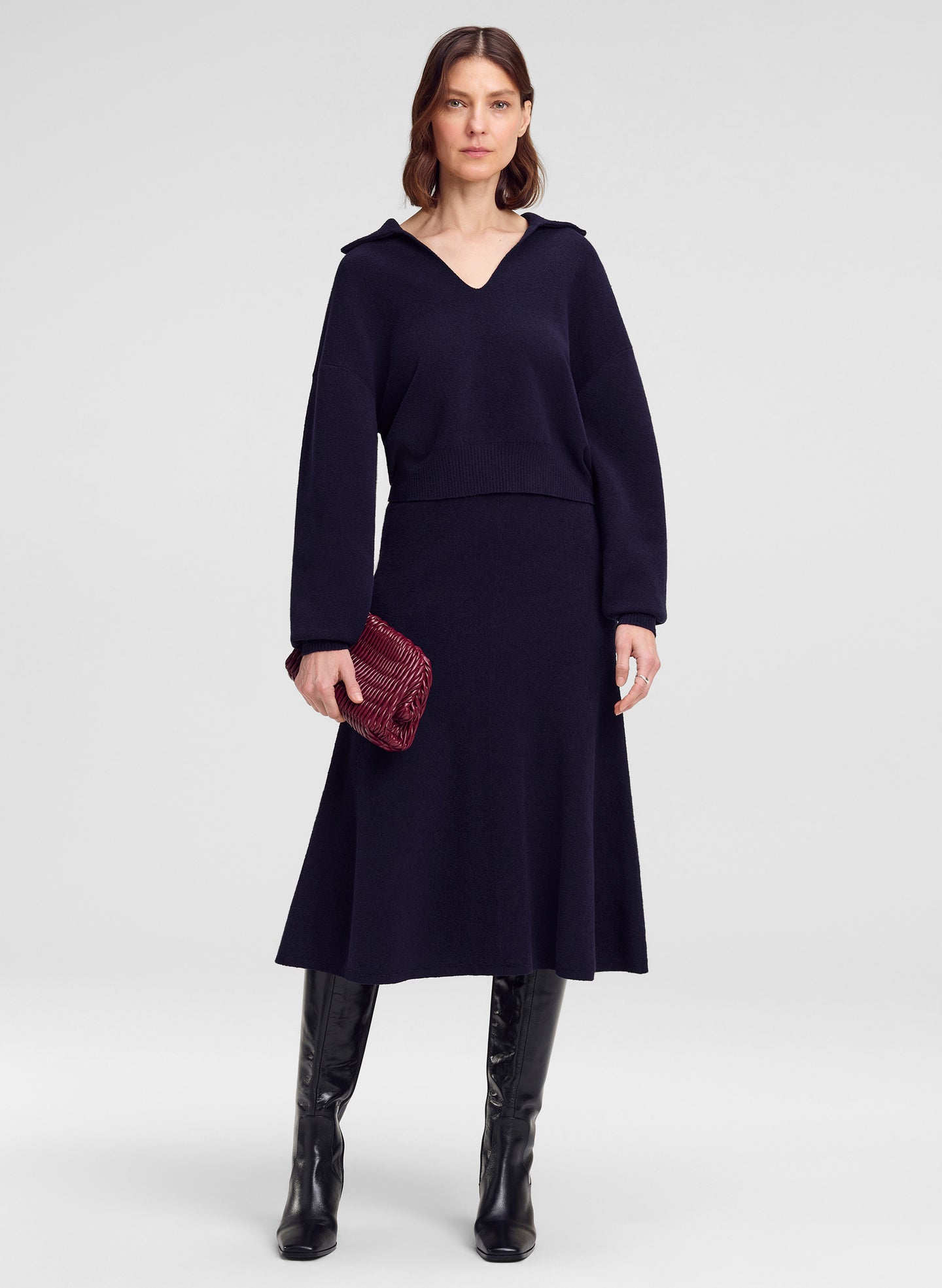 A model stands against a light gray studio background wearing a dark navy long-sleeve V-neck sweater with a flared skirt, paired with black knee-high boots and holding a small dark red textured clutch.