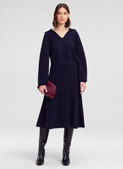 A model stands against a light gray studio background wearing a dark navy long-sleeve V-neck sweater with a flared skirt, paired with black knee-high boots and holding a small dark red textured clutch.