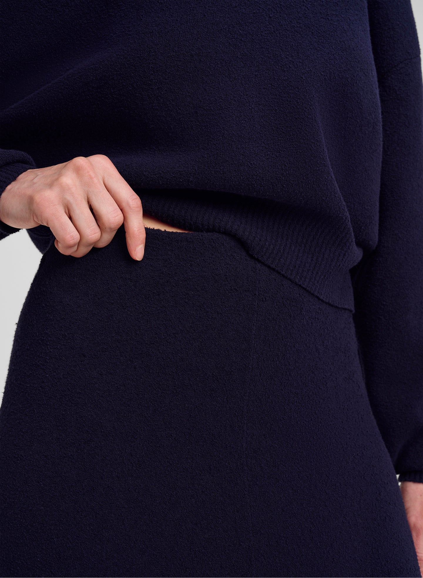 Close-up of a person wearing a dark navy knit sweater and matching skirt, with a hand pulling the hem of the sweater slightly to show the ribbed waistband.