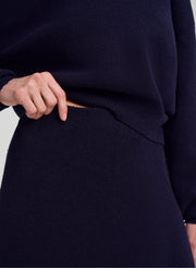 Close-up of a person wearing a dark navy knit sweater and matching skirt, with a hand pulling the hem of the sweater slightly to show the ribbed waistband.