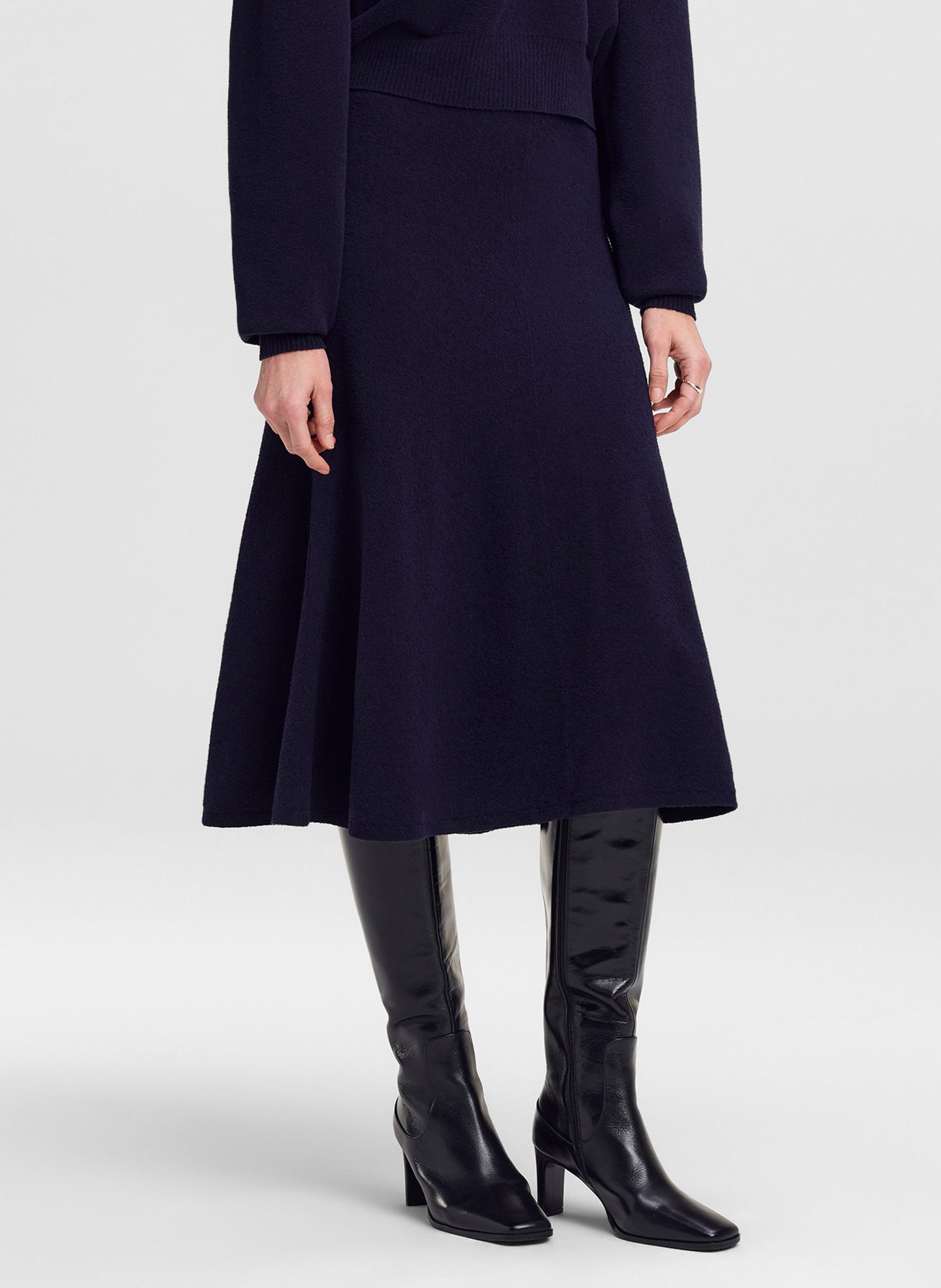 A model wearing a dark navy blue midi skirt with a flared A-line silhouette, paired with black leather knee-high boots, shown from the waist down on a light gray studio background.