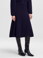 A model wearing a dark navy blue midi skirt with a flared A-line silhouette, paired with black leather knee-high boots, shown from the waist down on a light gray studio background.