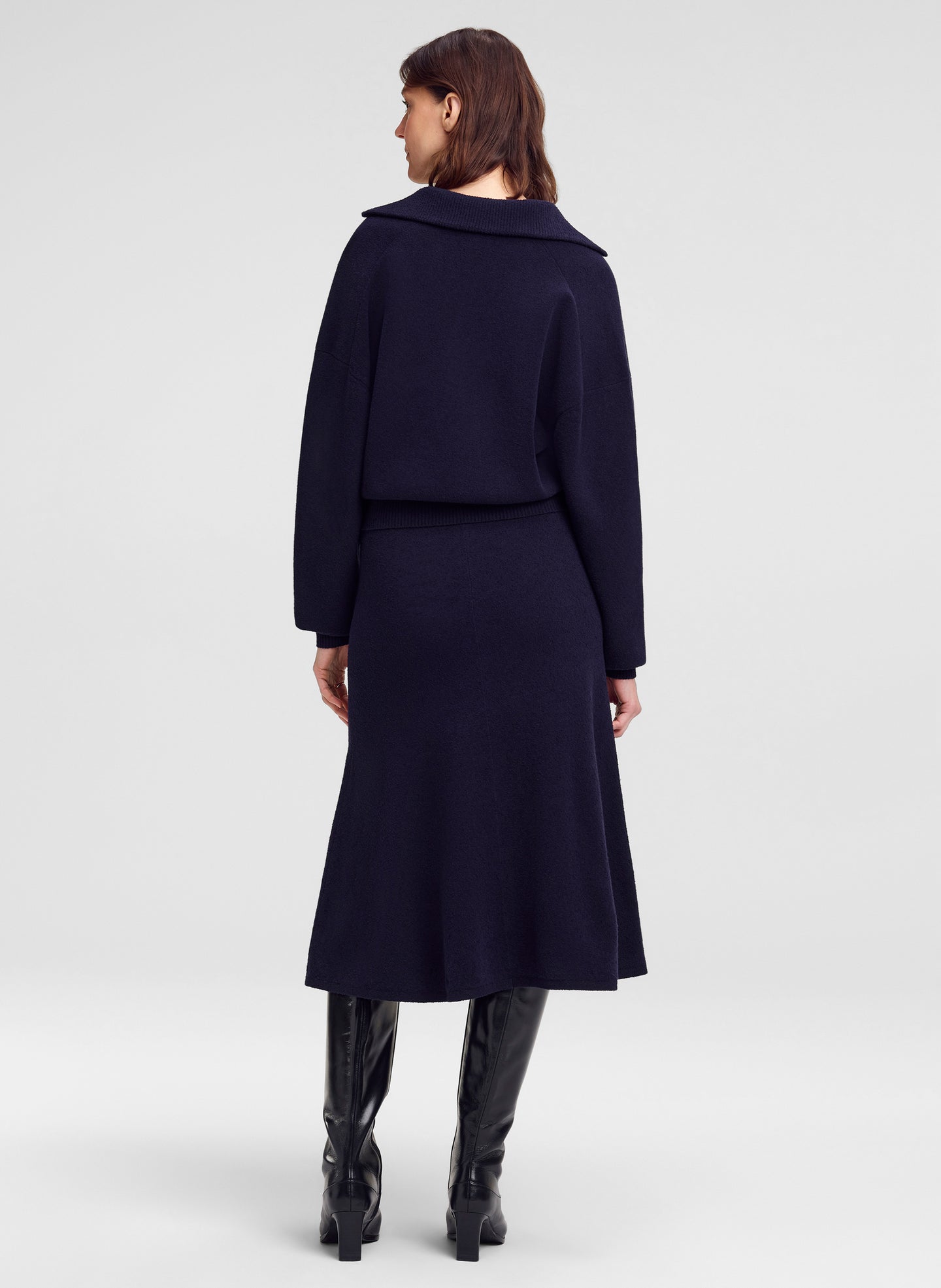 A woman shown from the back wearing a dark navy long-sleeve sweater with a wide collar and a matching midi skirt, paired with black knee-high boots, against a light gray studio background.