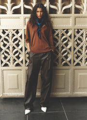 A model stands in front of an ornate beige metal gate, wearing a rust-brown long-sleeve top, dark brown leather trousers, white pointed-toe heels, and a blue neck scarf.