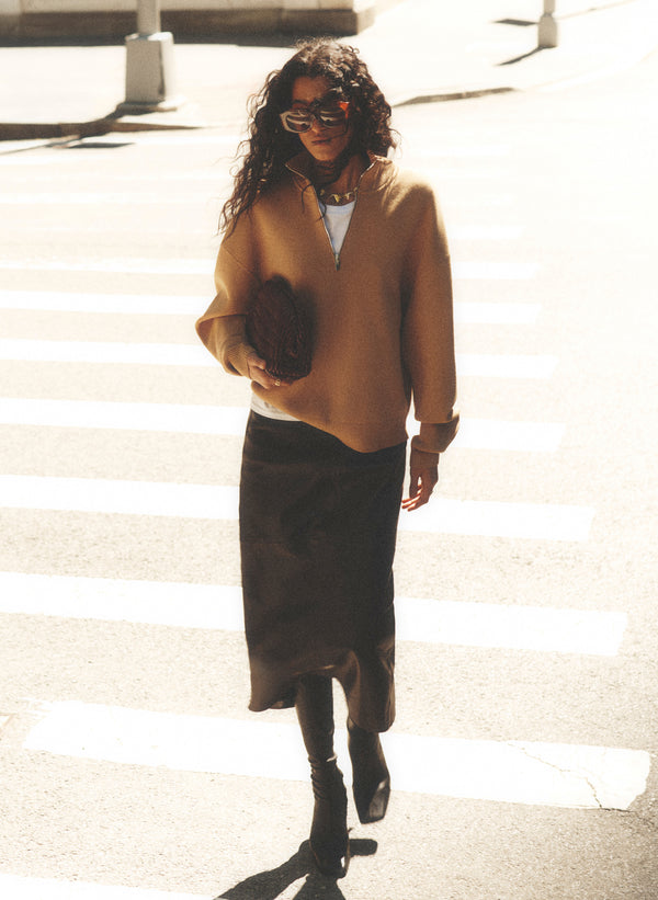 A woman walking on a sunlit city crosswalk wearing a camel-colored knit sweater, a dark midi skirt, black boots, and sunglasses.