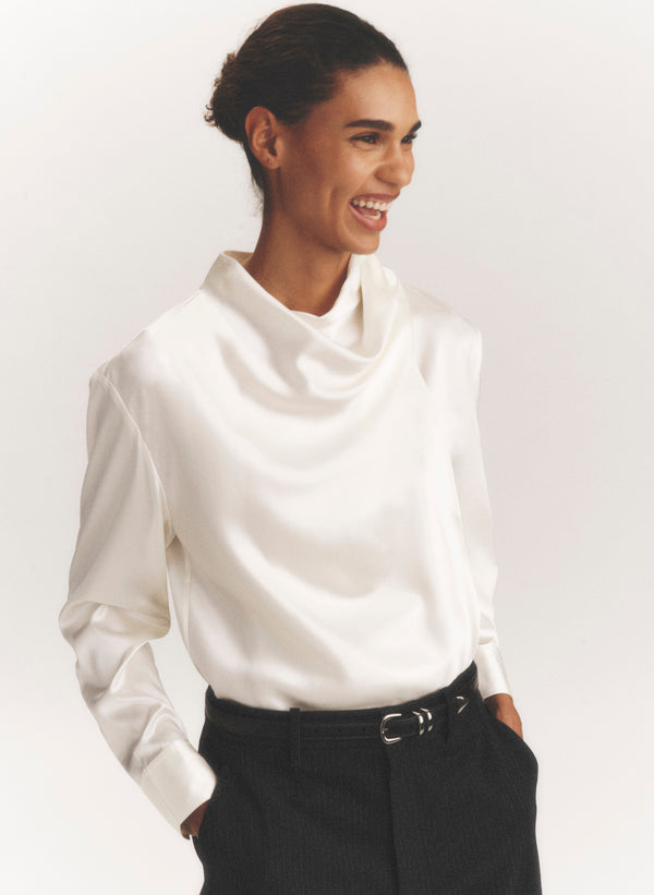 A woman wearing a white long-sleeve cowl-neck blouse tucked into black trousers, photographed against a clean light gray studio background.