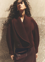 A woman with long, curly dark hair models a dark burgundy wrap-style coat and matching skirt against a textured concrete wall.
