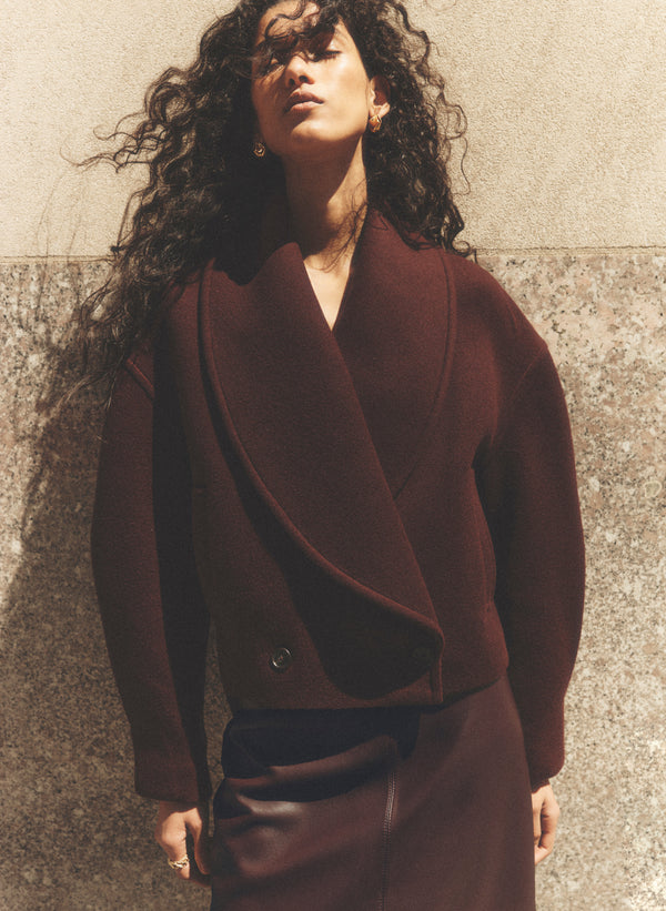 A woman with long, curly dark hair models a dark burgundy wrap-style coat and matching skirt against a textured concrete wall.