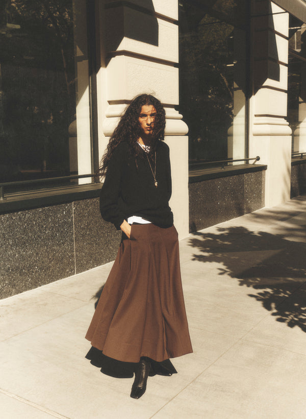A woman stands on a city sidewalk wearing a black long-sleeve top, a brown pleated maxi skirt, and black ankle boots, with her hands in her pockets.
