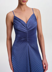 A woman wearing a blue pleated spaghetti-strap dress against a light gray studio background.