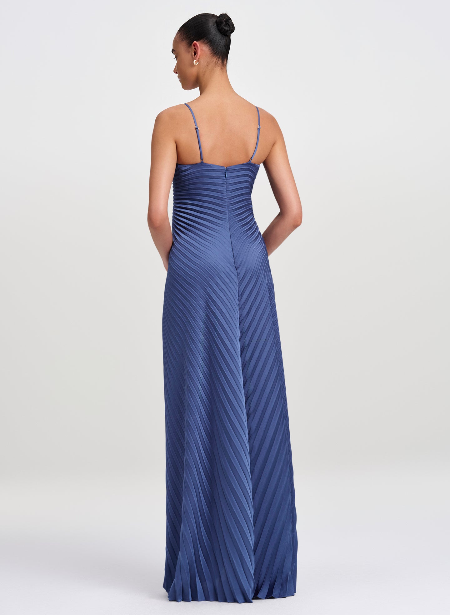 A woman modeling a floor-length pleated blue gown from the back, shown against a plain light gray studio background.
