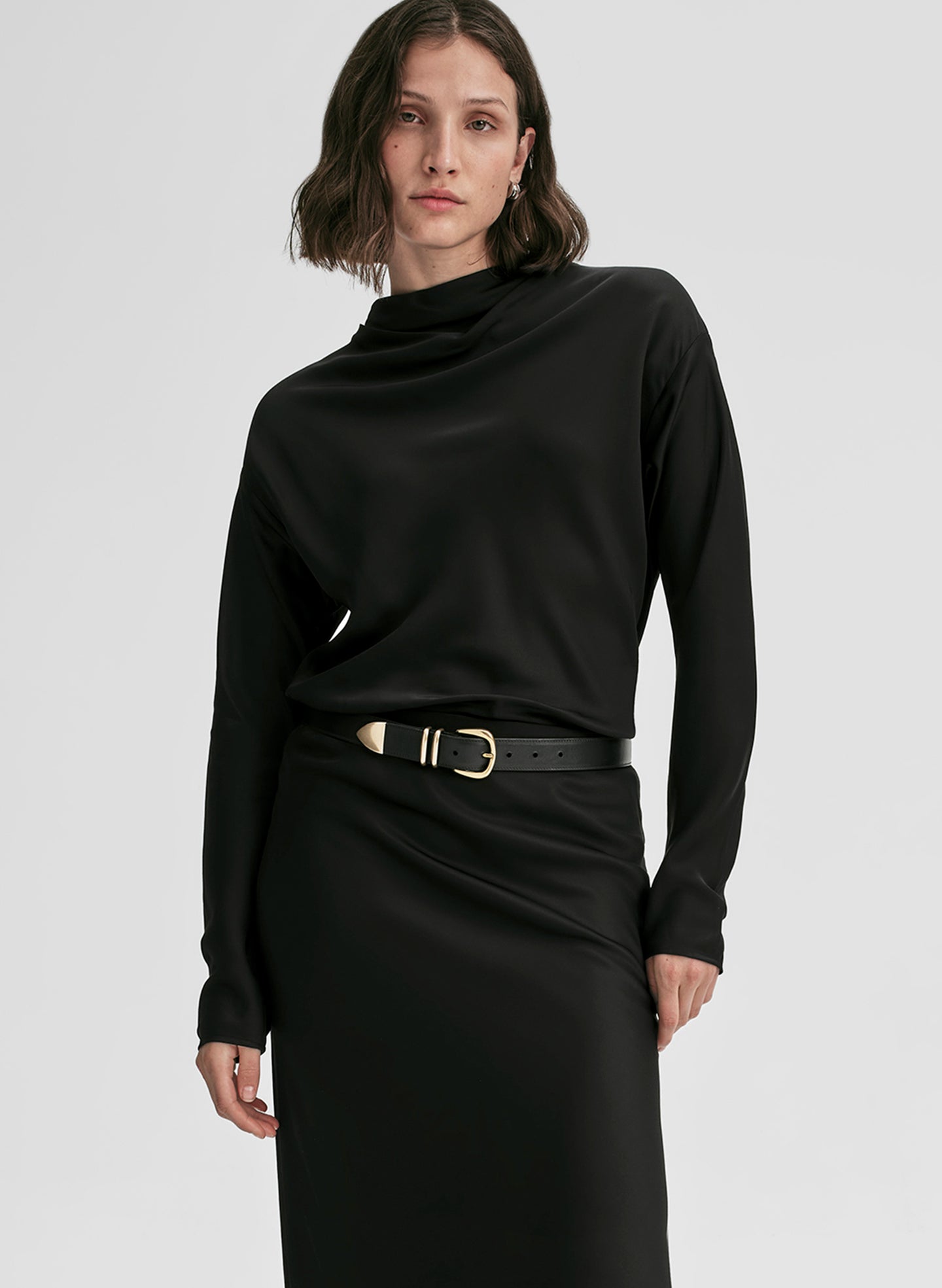 Woman wearing a black long-sleeve top and black skirt with a belt on a light gray background