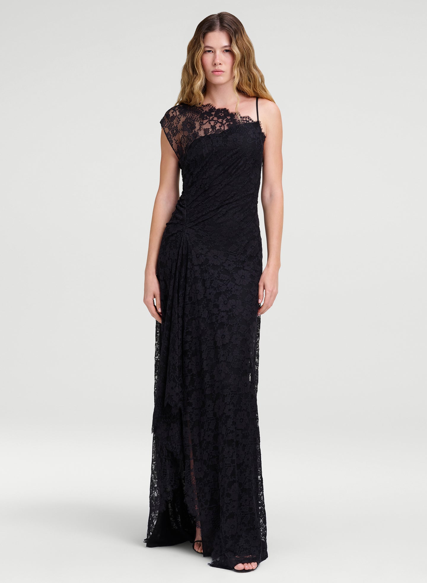 A woman modeling a floor-length black lace gown with thin straps, a draped one-shoulder neckline, and a gathered skirt, photographed against a light gray studio background.