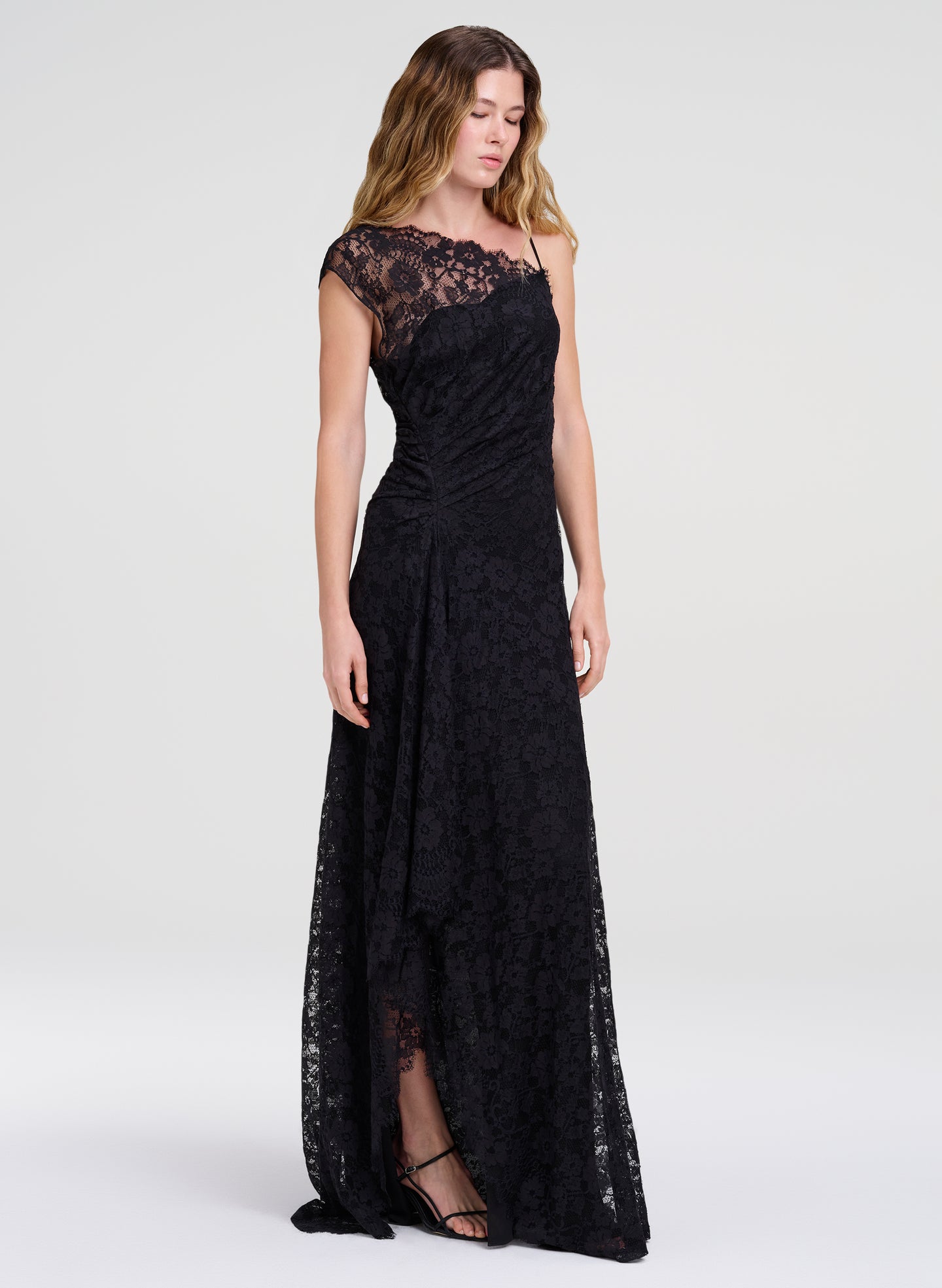 A woman modeling a floor-length black lace gown with cap sleeves and a draped neckline, photographed against a clean light gray studio background.